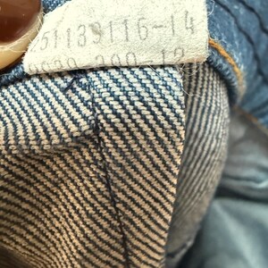 May include: Close-up of a blue denim waistband with a white tag that reads "251133116-14".