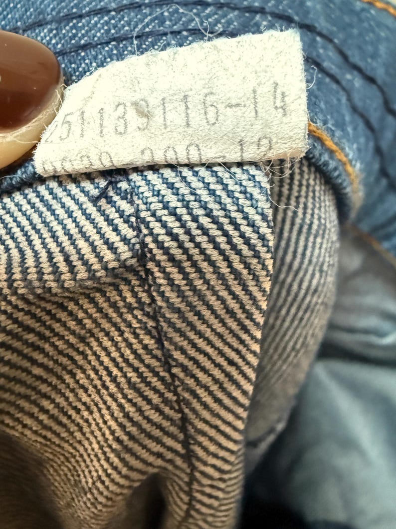 May include: Close-up of a blue denim waistband with a white tag that reads "251133116-14".