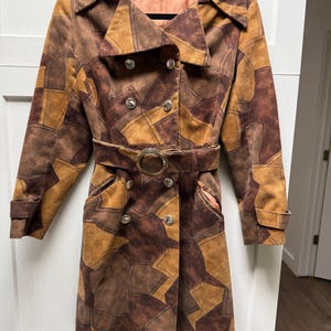 May include: A vintage patchwork coat in shades of brown and tan. The coat features a double-breasted closure with silver buttons, a matching belt with a circular buckle, and a collared neckline. The coat has a knee-length design.