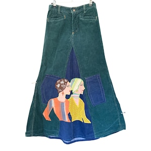 May include: A floor-length corduroy skirt in dark green featuring a central blue denim panel. The skirt includes two large blue denim pockets and a colourful applique graphic depicting two figures in profile, wearing patterned tops and headscarves.