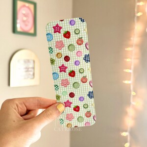 May include: A rectangular bookmark featuring a light green and white gingham pattern decorated with a repeating print of colorful buttons, star-shaped buttons, and small red strawberries scattered across the entire surface of the item.