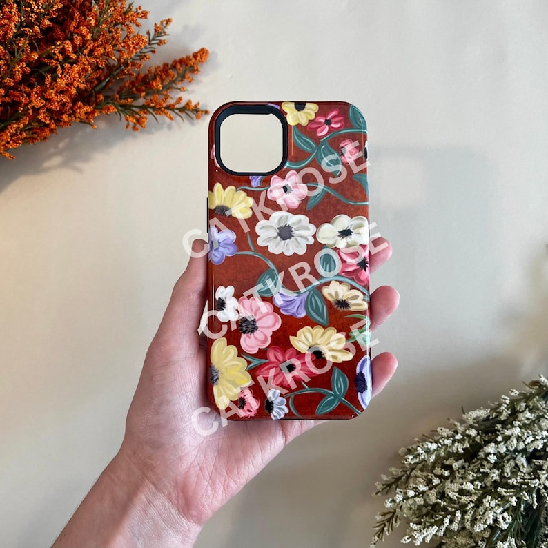 Op de afbeelding: Een rode telefoonhoes met een bloemenontwerp met roze, gele, witte en paarse bloemen. De bloemen zijn geschilderd op een donkerrode achtergrond met groene wijnstokken.