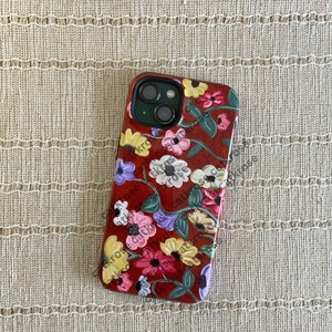 Op de afbeelding: Een rode telefoonhoes met een bloemenontwerp met roze, witte en gele bloemen. De hoes heeft een uitsparing voor de cameralens en een zwarte rand.
