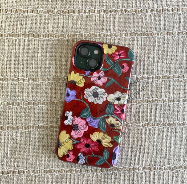 Op de afbeelding: Een rode telefoonhoes met een bloemenontwerp met roze, witte en gele bloemen. De hoes heeft een uitsparing voor de cameralens en een zwarte rand.
