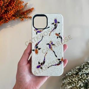 K&ouml;nnte beinhalten: Wei&szlig;e Handyh&uuml;lle mit glitzernder Oberfl&auml;che und einem Design aus bunten Koi-Fischen. Die H&uuml;lle hat einen Ausschnitt f&uuml;r die Kamera.