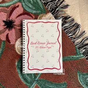 May include: A spiral-bound book review journal with a white cover featuring a red cherry border and scattered cherry illustrations. The text "Book Review Journal 25 Review Pages" is written in red script. The journal rests on a floral patterned blanket.
