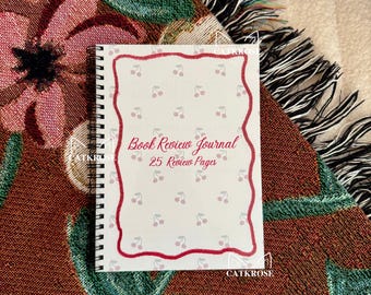 May include: A spiral-bound book review journal with a white cover featuring a red cherry border and scattered cherry illustrations. The text "Book Review Journal 25 Review Pages" is written in red script. The journal rests on a floral patterned blanket.