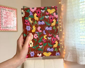 May include: A brown notebook with a floral pattern. The flowers are painted in shades of pink, purple, yellow, and white. The notebook is closed and held in a hand.