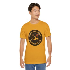 May include: A mustard yellow short-sleeve t-shirt featuring a circular black graphic on the chest. The graphic displays a mountain range, a planet with rings, and stars, with the text "NOVA EXPLORER ADVENTURE CO." printed in a circular border.