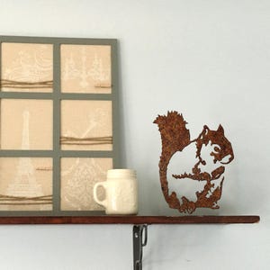 May include: A rustic metal squirrel silhouette in shades of brown, perched on a wooden shelf. The squirrel is the focal point, with a detailed cutout design. A framed artwork with beige and white prints and a small white mug are also on the shelf.