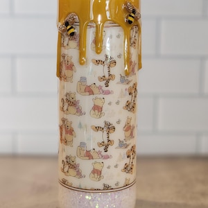May include: A white tumbler with a Winnie the Pooh and Tigger pattern. The tumbler has a gold glitter bottom and a yellow honey drip design with bee accents.