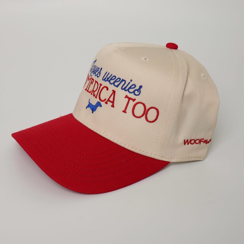 May include: Cream-colored baseball cap with a red brim and top button. The cap features embroidered text in blue and red that reads "Loves weenies AMERICA TOO" with a blue dachshund graphic. The word "WOOF" is embroidered on the side.