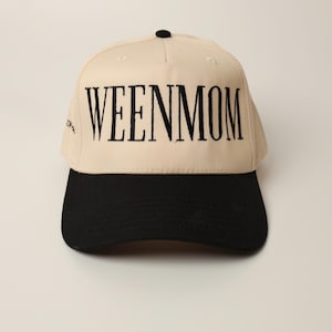 May include: A beige baseball cap with a black brim and a black button on top. The cap features the embroidered text "WEENMOM" in large, black capital letters across the front.