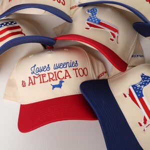 May include: A collection of cream-colored baseball caps with red and blue accents. Some caps feature an American flag design in the shape of a dachshund, while others display the text "Loves weenies & AMERICA TOO".