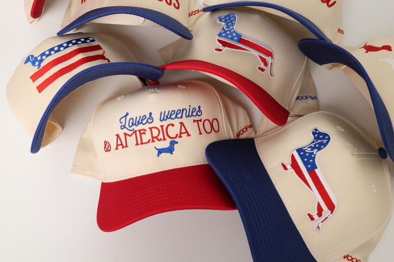 May include: A collection of cream-colored baseball caps with red and blue accents. Some caps feature an American flag design in the shape of a dachshund, while others display the text "Loves weenies & AMERICA TOO".