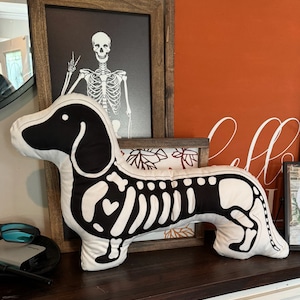 May include: A white plush dachshund pillow with a black skeleton design. The pillow is lying on a dark brown surface with a wooden frame in the background.