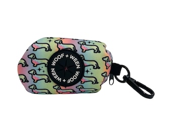 May include: A rainbow-colored waste bag dispenser featuring a repeating pattern of dachshund silhouettes and small stars. The pouch has a black circular rubber grommet in the center with the white text WOOF + WEEN printed twice and a black clip.
