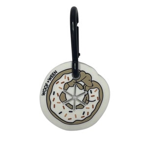 May include: White donut-shaped dog treat holder with a black carabiner clip. The holder has a brown and white dog illustration inside the donut and the text "WOOF + WEEN" on the side.
