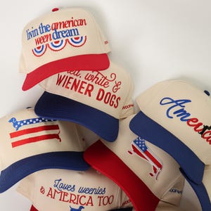 May include: A collection of cream-colored baseball caps with red and blue accents. Each cap features embroidered text and graphics, including phrases like "Livin the American Ween Dream" and "America." The caps have a patriotic theme.