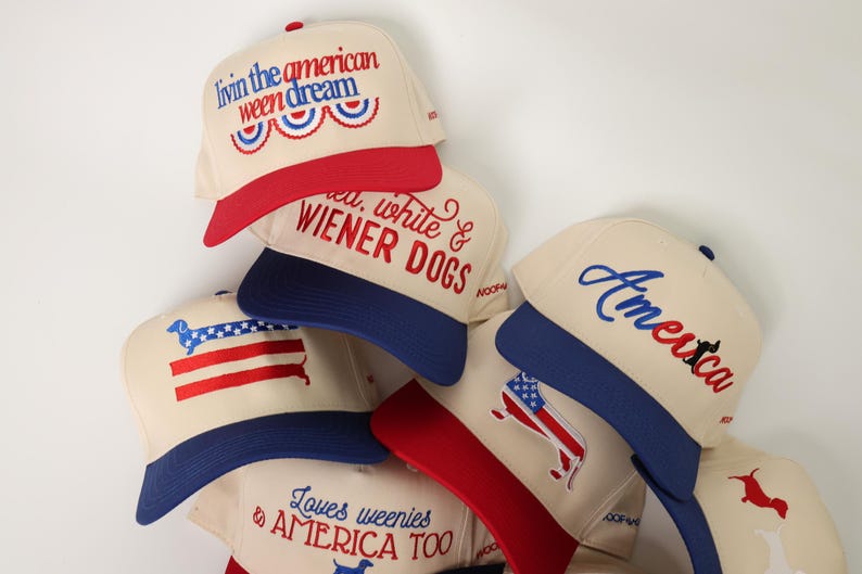 May include: A collection of cream-colored baseball caps with red and blue accents. Each cap features embroidered text and graphics, including phrases like "Livin the American Ween Dream" and "America." The caps have a patriotic theme.