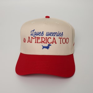 May include: A baseball cap with a cream-colored crown and a red brim. The cap features embroidered text in blue and red that reads "Loves weenies & AMERICA TOO" with a blue dachshund silhouette.