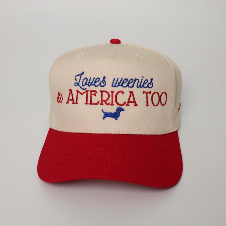 May include: A baseball cap with a cream-colored crown and a red brim. The cap features embroidered text in blue and red that reads "Loves weenies & AMERICA TOO" with a blue dachshund silhouette.