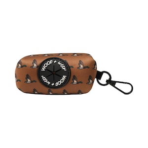 May include: Brown dog waste bag dispenser with a black carabiner clip. The dispenser is covered in a pattern of flying ducks and features the text "WOOF + ween" around a central opening.