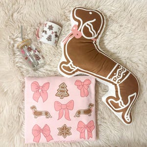 May include: A collection of holiday-themed items, including a gingerbread dachshund pillow, a pink graphic tee with bows and dachshunds, a glass tumbler, and a mug. The items are arranged on a white fluffy surface.