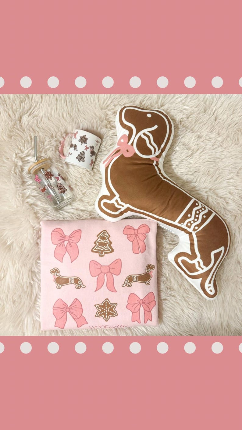 May include: A collection of holiday-themed items, including a gingerbread dachshund pillow, a pink graphic tee with bows and dachshunds, a glass tumbler, and a mug. The items are arranged on a white fluffy surface.