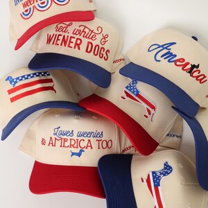 May include: A collection of cream-colored baseball caps with red and blue accents. Each cap features patriotic text and dachshund dog graphics. The phrases include "Livin' the American Ween Dream" and "Red, White & Wiener Dogs."