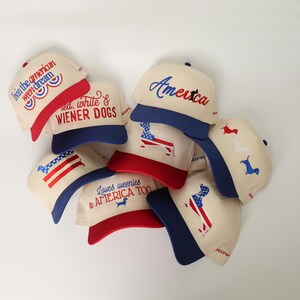 May include: A collection of cream-colored baseball caps with red and blue accents. Each cap features patriotic designs, including the American flag, the word "America," and the phrase "Loves weenies & AMERICA TOO."