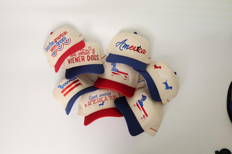 May include: A collection of cream-colored baseball caps with red and blue accents. Each cap features patriotic designs, including the American flag, the word "America," and the phrase "Loves weenies & AMERICA TOO."