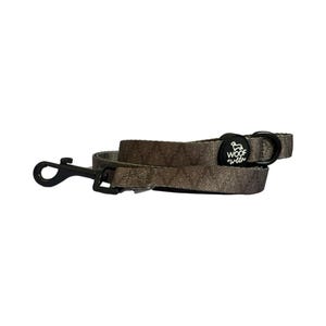 May include: A dog leash with a repeating leaf pattern in shades of brown and tan. The leash has a black metal clasp and a black plastic buckle. The leash has the words "WOOF + WELL" printed on a black circular patch.