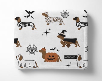 May include: White fleece blanket with a repeating pattern of dachshund dogs dressed in various Halloween costumes, including a skeleton, a mummy, a witch, a ghost, and a pumpkin. The blanket also features bats, stars, and spiderwebs. The text "WOOF+WIM" is printed in the bottom right corner.
