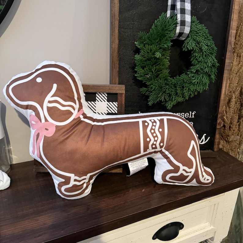 May include: A brown and white dachshund-shaped pillow with a pink bow. The pillow has a gingerbread cookie design with white icing details. The pillow is on a dark wooden surface.