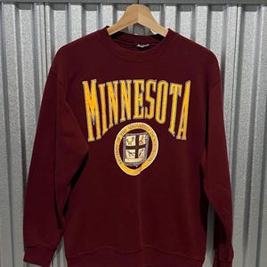 May include: Maroon crewneck sweatshirt with the word "MINNESOTA" in gold lettering across the chest. Below the text is a circular emblem with the words "THE UNIVERSITY OF MINNESOTA" and a crest. The sweatshirt is hanging on a wooden hanger.