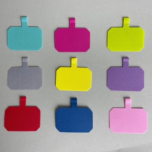 Può includere: Nove etichette in tessuto colorate con un anello in cima. Le etichette sono nei seguenti colori: azzurro, rosa, giallo, grigio, giallo, viola, rosso, blu e rosa.