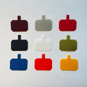 Peut inclure: Une collection de neuf étiquettes rectangulaires en tissu avec des coins arrondis et une petite languette verticale sur le dessus. Les étiquettes sont disposées en grille de trois par trois et présentent des couleurs unies : bordeaux, gris, rouge, noir, blanc, vert olive, bleu, rouge vif et jaune.