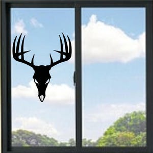 Peut inclure: Silhouette noire d'un crâne de cerf avec de grands bois sur une fenêtre. La fenêtre encadre un ciel bleu nuageux et des arbres verts. Le crâne de cerf est un élément décoratif.