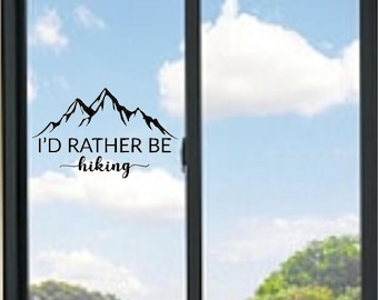 May include: A black vinyl decal applied to a window pane, featuring a stylized mountain range silhouette above the text I'D RATHER BE hiking in a combination of bold sans-serif and elegant cursive script lettering for a clear visual display.