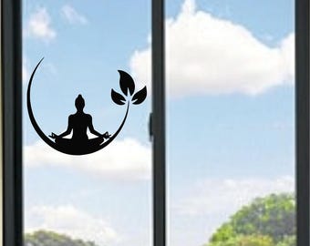 Puede incluir: Un adhesivo de vinilo negro aplicado sobre un cristal de ventana. El diseño presenta la silueta de una persona en posición de meditación sentada, situada dentro de una forma de media luna que se extiende en un tallo con tres hojas.