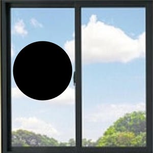 May include: A black circular decal applied to the left pane of a dark-framed sliding window. The window features two glass panels set within a rectangular frame, with a view of a blue sky, white clouds, and green trees visible through the glass.