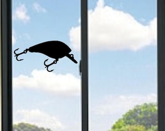 May include: A black silhouette decal of a fishing lure featuring a rounded body shape, a small eye detail, and two treble hooks attached to the underside and rear. The graphic is applied to a glass window pane with a dark frame.