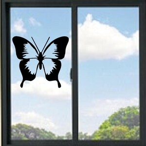 Può includere: Un adesivo a forma di farfalla in bianco e nero è applicato a una finestra. La farfalla presenta un motivo alare dettagliato, sullo sfondo di un cielo blu con nuvole e alberi verdi.