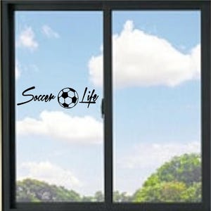 May include: A black vinyl decal applied to a glass window pane. The decal features the text Soccer in a cursive script, followed by a graphic of a football, and the word Life in a cursive script, all in a solid black colour.