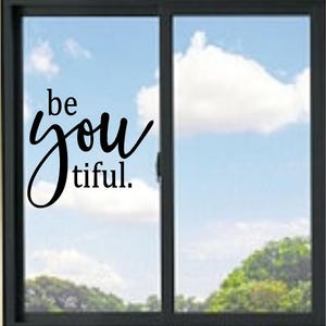 May include: A black vinyl window decal applied to a glass pane, featuring the text "be you tiful." The word "be" and "tiful." are in a simple serif font, while the word "you" is written in a large, flowing, cursive script style.