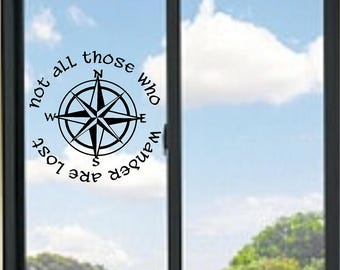 Puede incluir: Un adhesivo de vinilo negro aplicado sobre un cristal de ventana. El diseño presenta una rosa de los vientos central con los puntos cardinales N, S, E y O, rodeada por el texto circular "not all those who wander are lost" en una fuente estilizada.