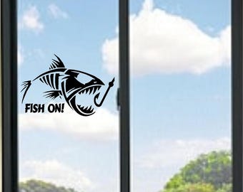 May include: A black vinyl decal applied to a glass window, featuring a stylized fish skeleton graphic with an open mouth and a fishing hook. The text FISH ON! is printed in a bold, sans-serif font directly below the fish skeleton design.