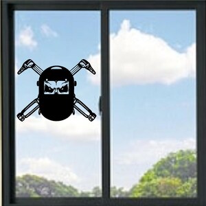 May include: A black vinyl decal depicting a welder's mask with crossed welding torches. The mask has an angry face design. The decal is displayed on a window with a blue sky and clouds in the background.
