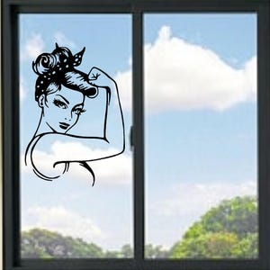 May include: A black and white graphic of a woman flexing her bicep, a symbol of strength and empowerment. The woman has her hair up in a bun with a polka dot scarf. The image is on a window with a blue sky and clouds in the background.